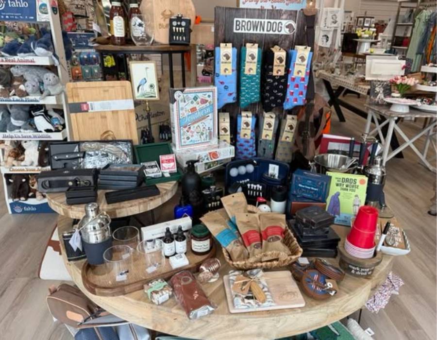 Gift display at Lettrs Gifts & Embroidery in Bluffton, SC, featuring personalized men’s gifts, socks, barware, skincare products, and novelty items on rustic wooden tables inside a boutique gift shop.