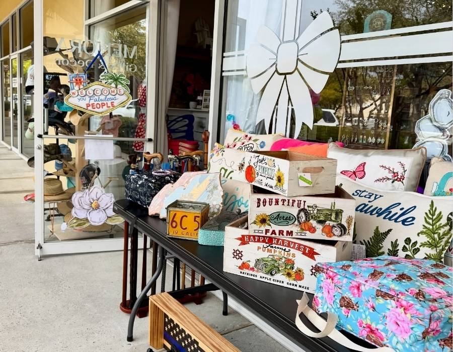 Colorful outdoor display at Meliora Gifts & Decor in Bluffton, SC, featuring decorative throw pillows, rustic harvest crates, floral bags, novelty signs, and unique home accessories outside a boutique storefront.