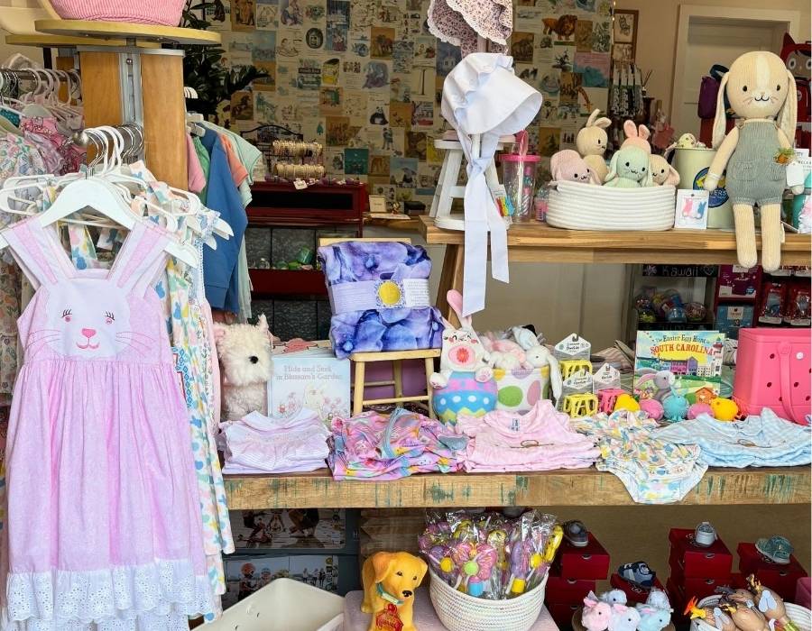 Colorful baby boutique display at Moonlit Lullaby gift shop in Bluffton, SC, featuring pastel baby clothes, stuffed animals, handmade dolls, and children’s books arranged on rustic wood shelves.